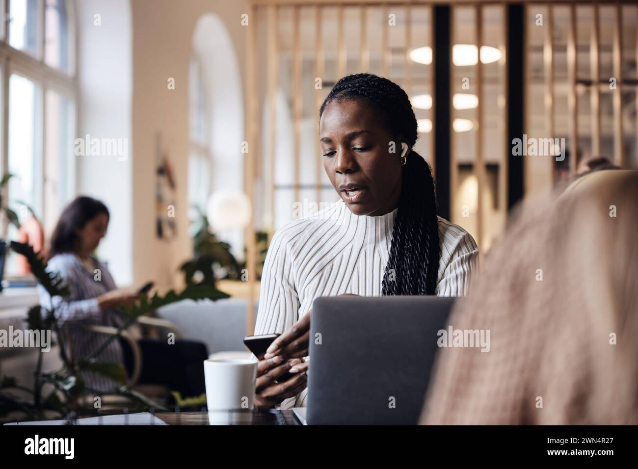 Femme d'affaires parlant à travers des écouteurs intra-auriculaires sans fil et à l'aide d'un téléphone intelligent tout en étant assis au bureau de coworking Banque D'Images