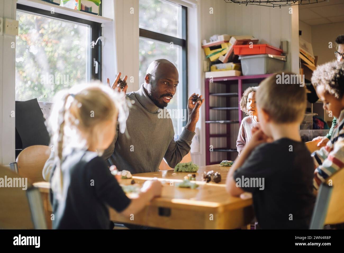 Enseignant masculin expliquant aux enfants assis sur le banc pendant le cours de moulage d'argile à la maternelle Banque D'Images
