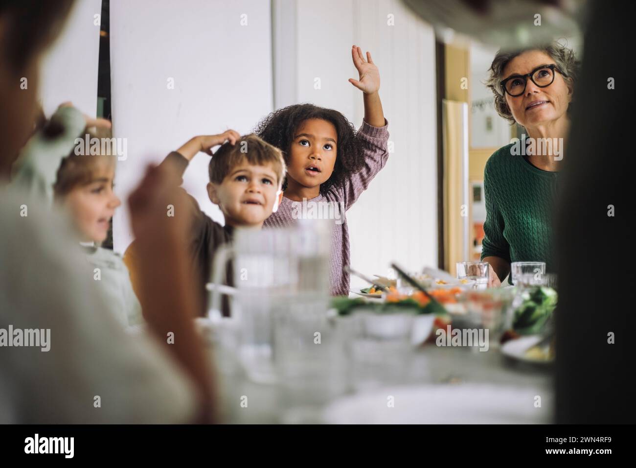 Les enfants levant la main tout en étant assis avec une enseignante pendant l'heure du déjeuner à la maternelle Banque D'Images