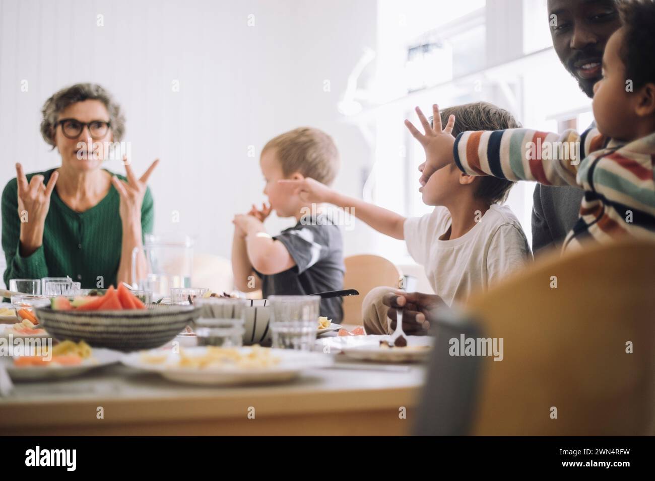Les élèves d'âge préscolaire faisant des gestes à l'enseignant tout en étant assis à la table à manger pendant la pause déjeuner Banque D'Images