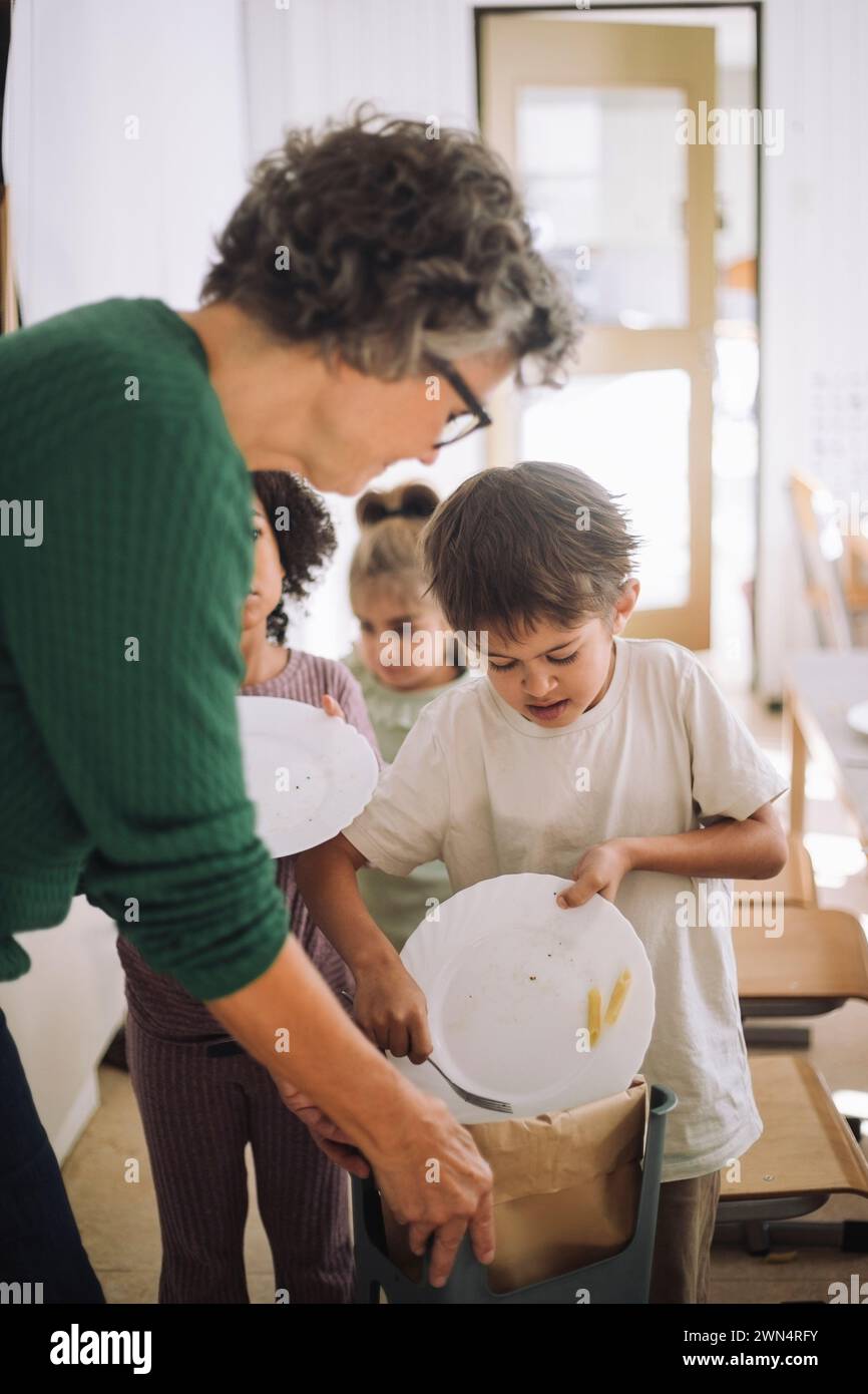 Les enfants jetant des restes à la poubelle peuvent être gardés par le professeur à la maternelle Banque D'Images