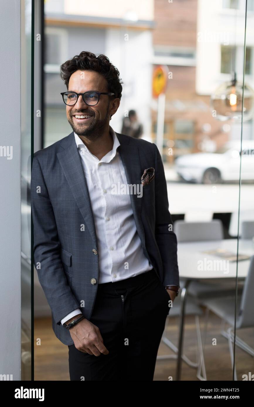 Agent immobilier masculin souriant appuyé sur la porte vitrée dans le bureau Banque D'Images