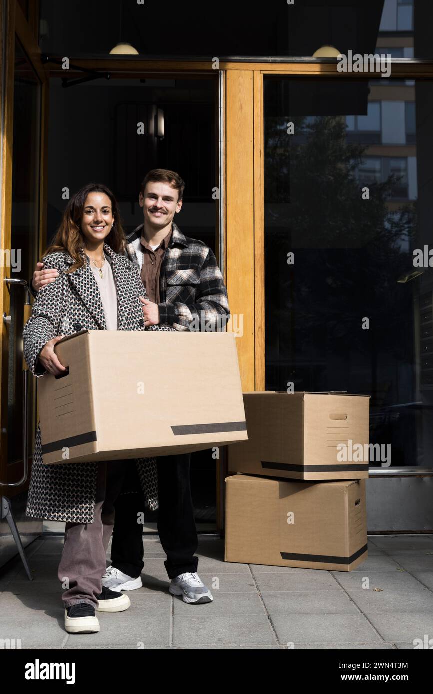 Portrait de couple multiracial souriant debout avec des boîtes en face de l'appartement Banque D'Images