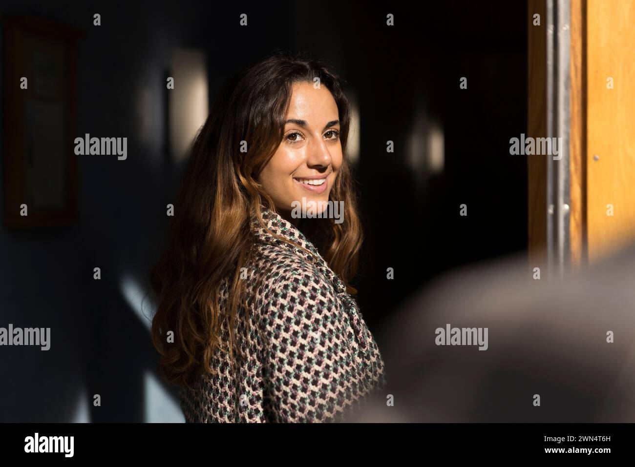 Femme souriante avec de longs cheveux regardant par-dessus l'épaule à l'entrée de l'appartement Banque D'Images