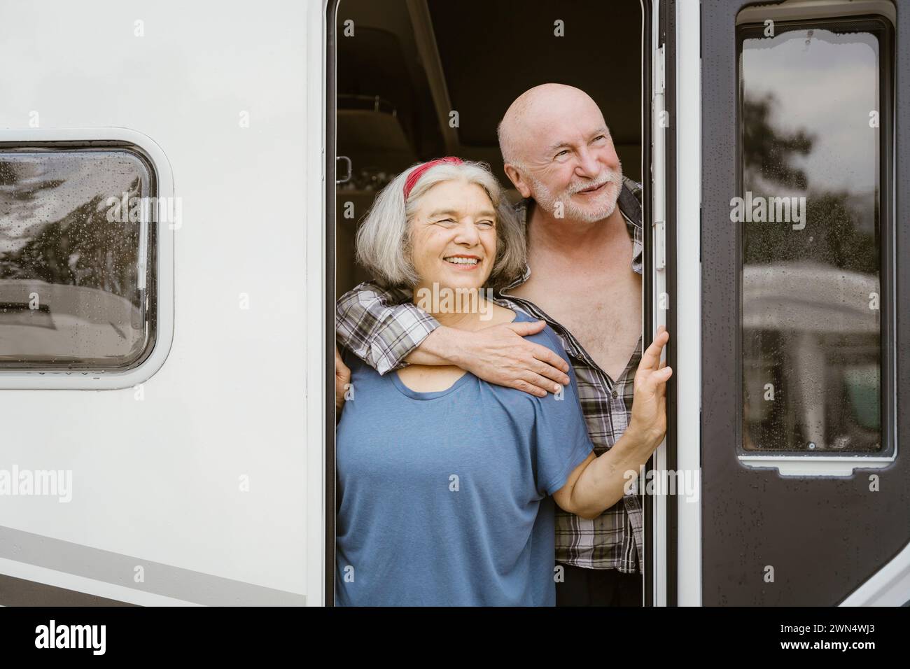 Homme senior souriant embrassant la femme par derrière tout en se tenant à la porte du camping-car Banque D'Images