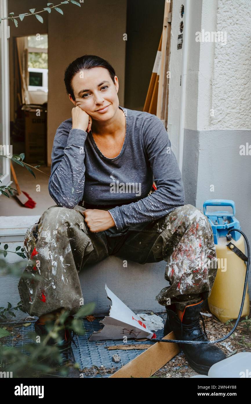 Portrait pleine longueur de charpentier féminin souriant assis à la porte de la maison Banque D'Images