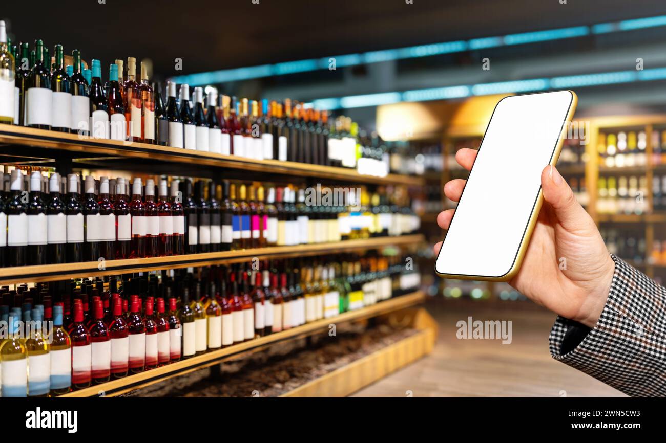 Main féminine montrant l'écran vide de son téléphone intelligent devant le magasin d'alcool avec des bouteilles de vin sur les étagères. Banque D'Images