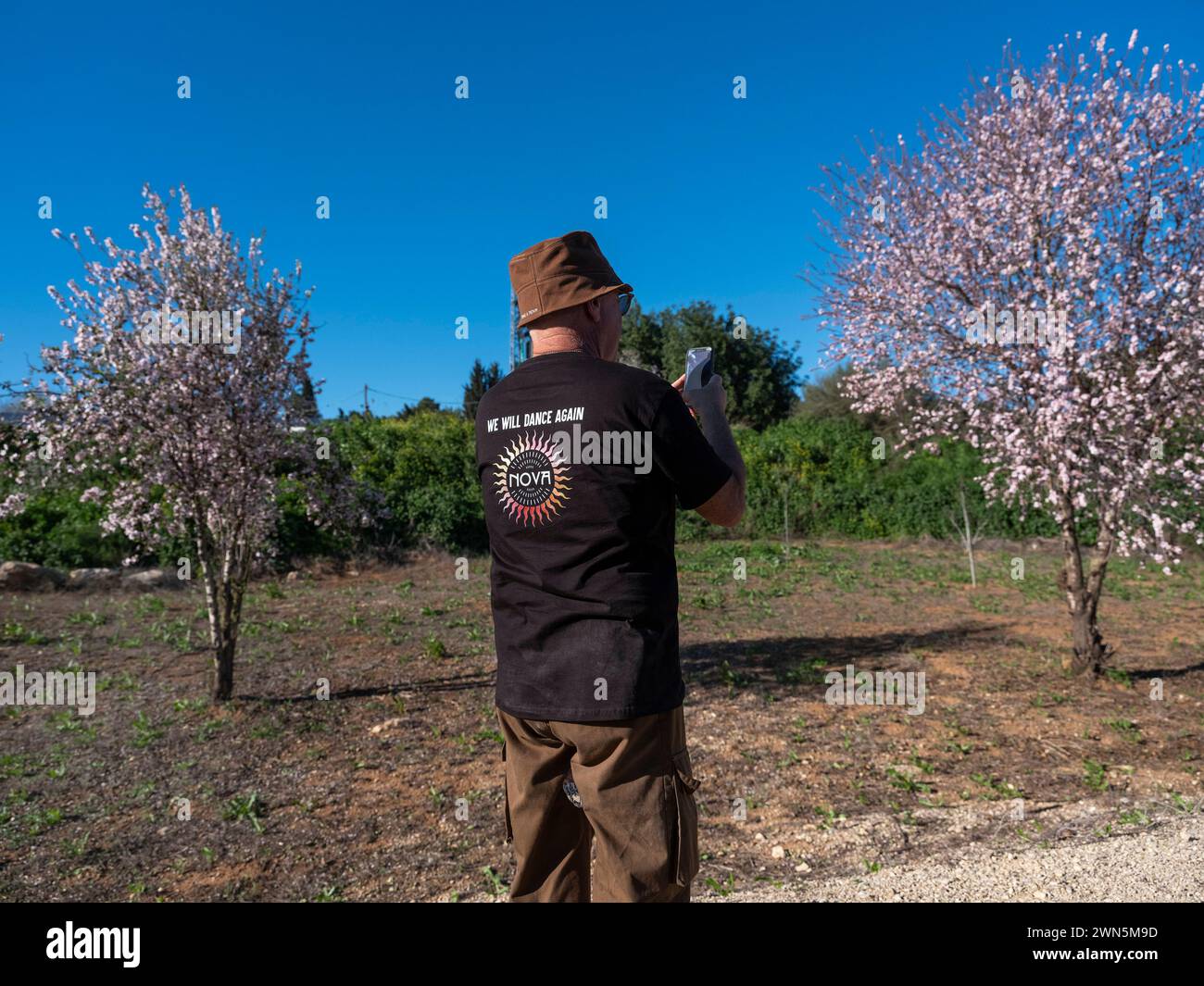 Un israélien s’arrête pour prendre une photo des arbres fruitiers en fleurs à l’entrée de Beit Guvrin dans le centre d’Israël le 29 février, 2024 alors qu'il porte un tee-shirt disant "nous danserons à nouveau Nova" alors que quelque 500 marcheurs et membres des familles d'otages détenus dans la bande de Gaza marchent vers Jérusalem. Quelque 500 Israéliens défilent aujourd’hui le deuxième jour de leur marche depuis les communautés frontalières de la bande de Gaza vers Jérusalem, appelant le gouvernement à prendre la décision de libérer les otages détenus depuis la frénésie meurtrière du Hamas le 7 octobre 2023, il y a plus de quatre mois complets. Photo de Jim Hollander/UPI Banque D'Images