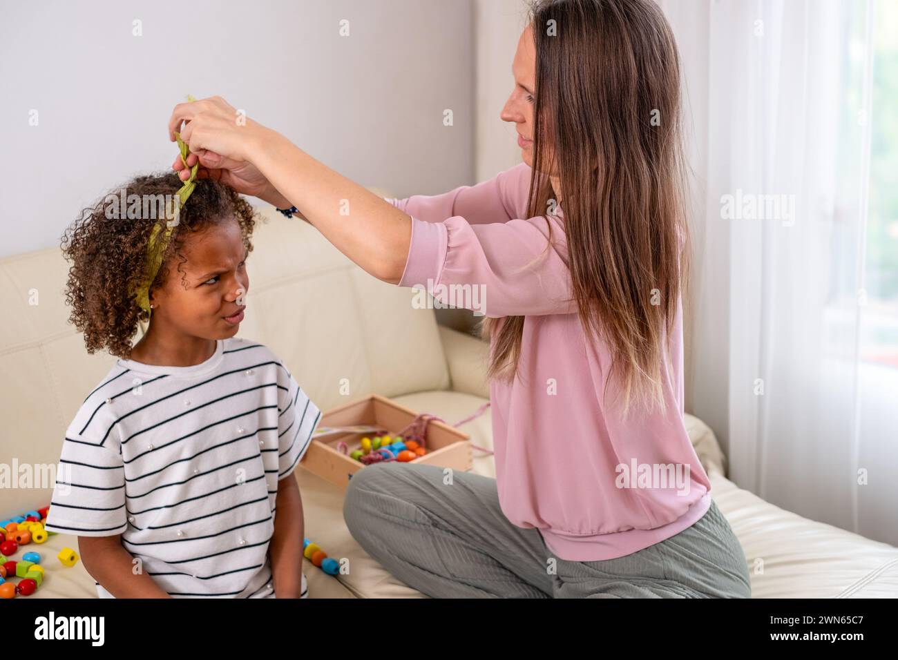 Une mère attache tendrement ses cheveux de filles confondus de race mixte, tous deux souriants, dans une pièce confortable et ensoleillée un moment d'amour maternel. Photo de haute qualité Banque D'Images