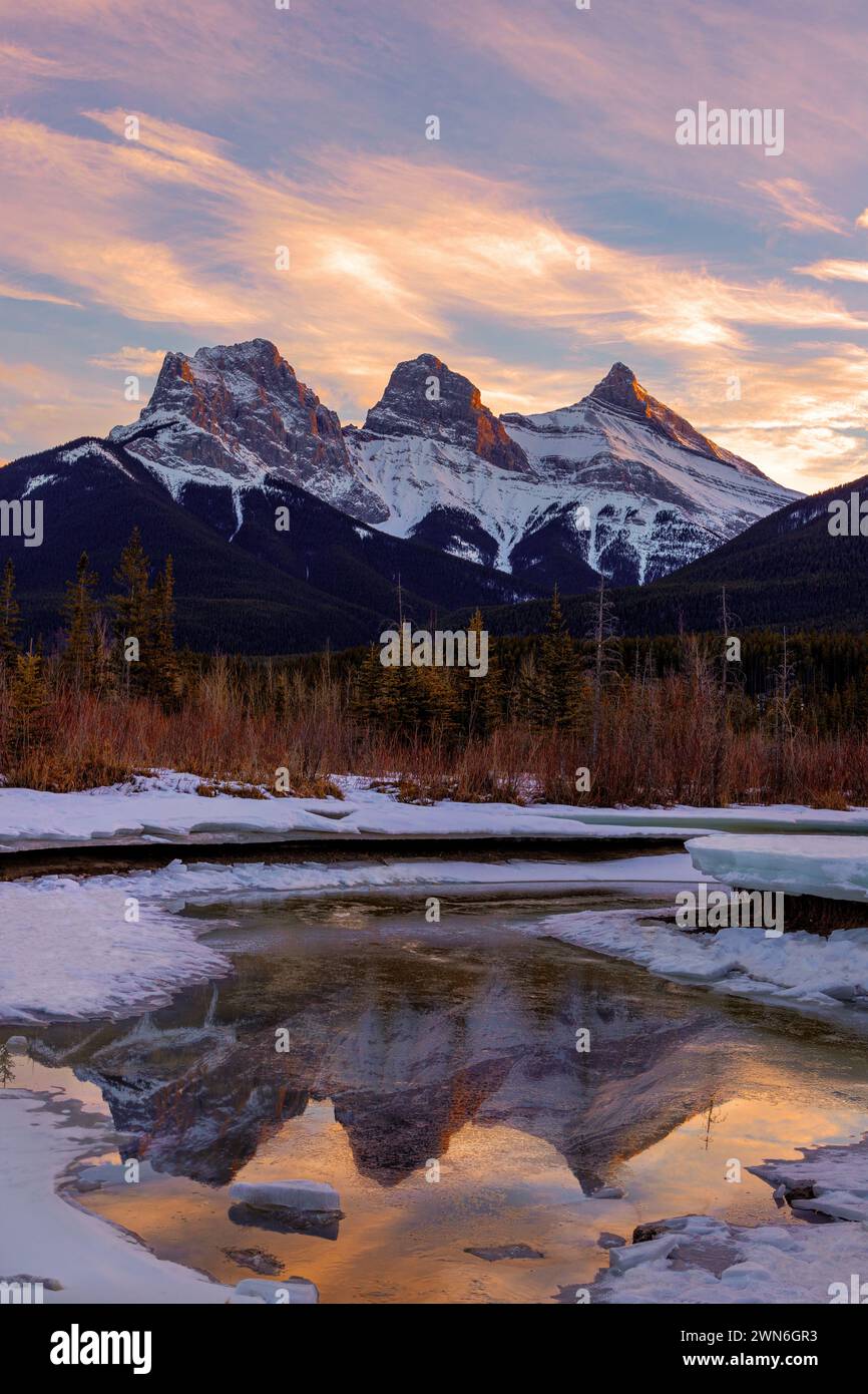 Coucher de soleil doré d'hiver aux trois Sœurs, un trio de sommets près de Canmore, Alberta, Canada, avec alpenglow sur les montagnes reflétant sur la glace dans le fo Banque D'Images