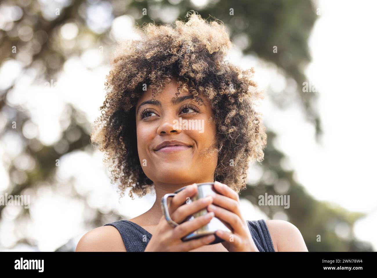 Une jeune femme biraciale aux cheveux bouclés tient une tasse à l'extérieur Banque D'Images