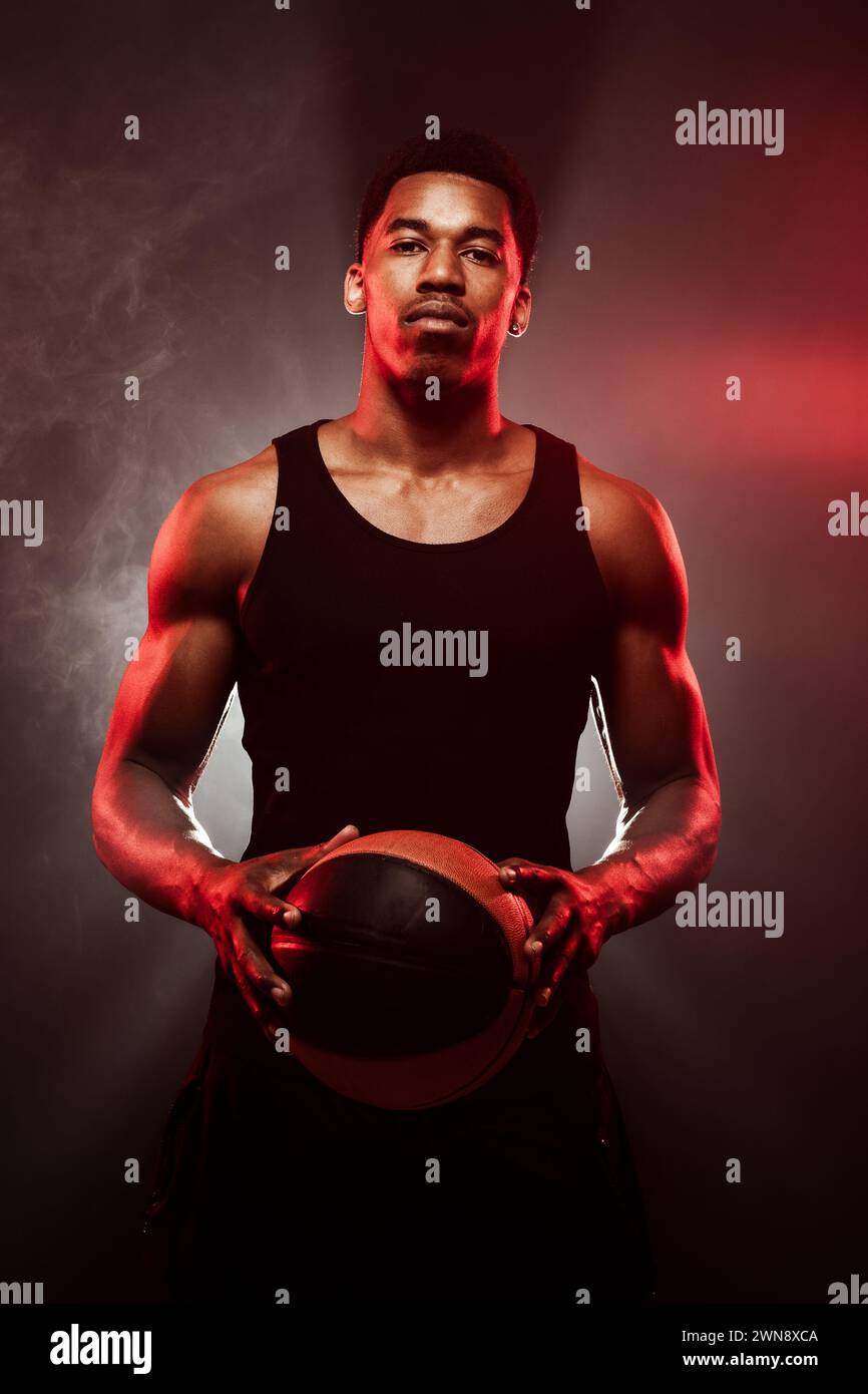 Côté joueur de basket-ball éclairé avec une couleur rouge tenant une balle sur fond de brouillard de fumée brumeux. Homme afro-américain concentré sérieux. Banque D'Images