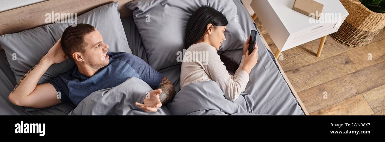homme irrité se querellant contre la jeune femme asiatique regardant dans son smartphone dans la chambre, bannière Banque D'Images