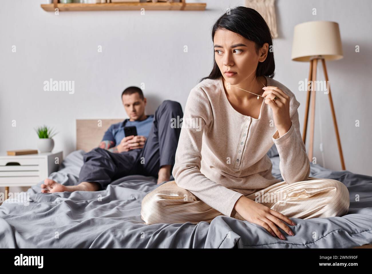 femme asiatique bouleversée assise près du mari à l'aide d'un smartphone dans la chambre à coucher à la maison, problème familial Banque D'Images