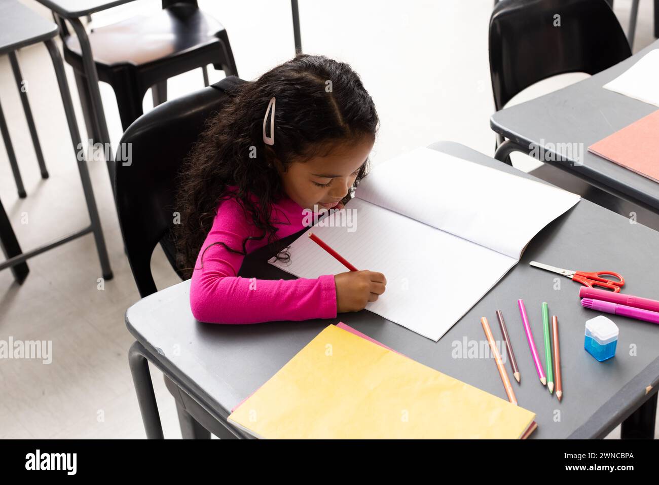 Fille biraciale avec les cheveux bouclés écrit dans un cahier à un bureau de classe d'école Banque D'Images