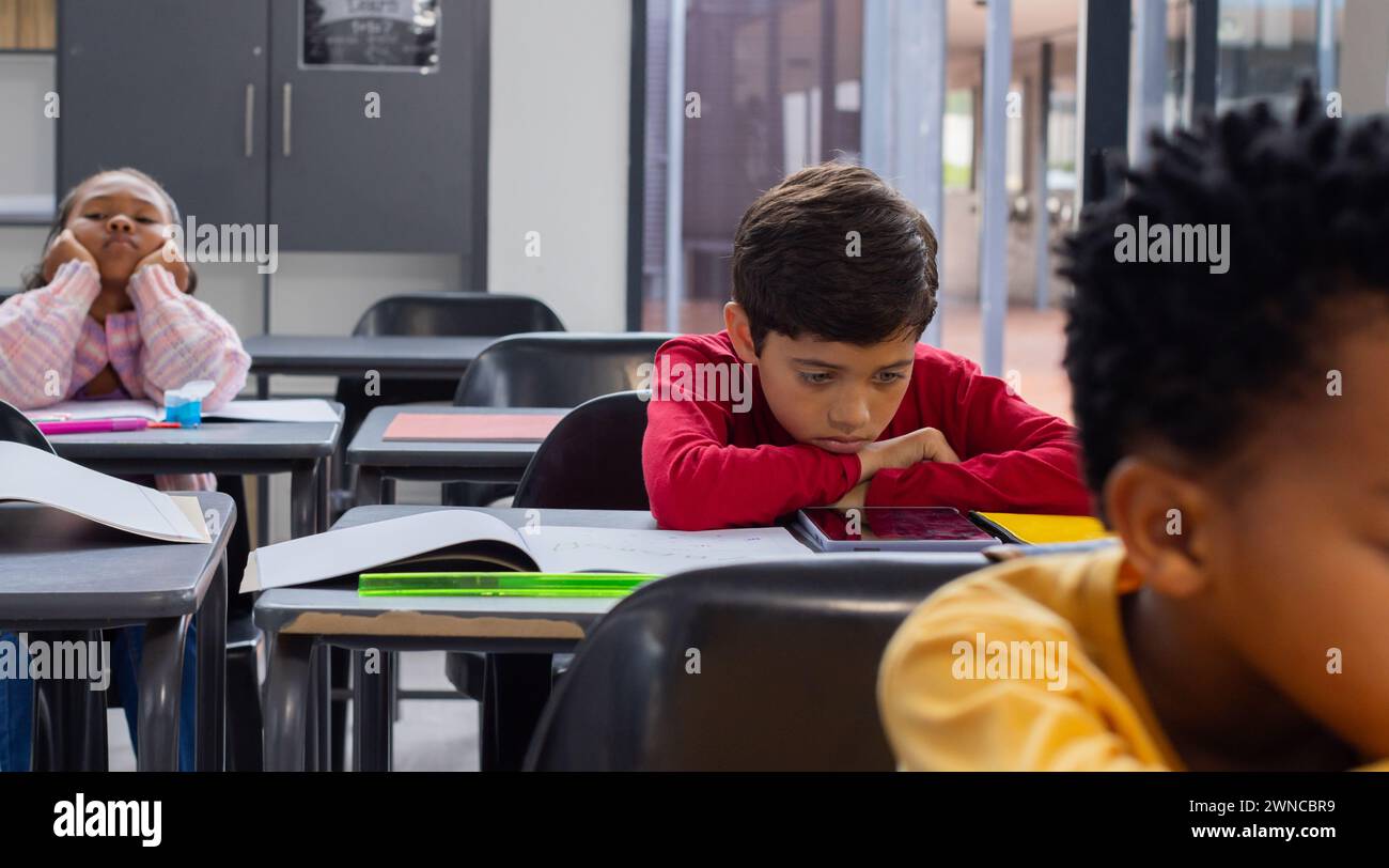 Garçon biracial en rouge semble triste à son bureau dans une salle de classe d'école, avec un espace de copie, camarades de classe autour Banque D'Images