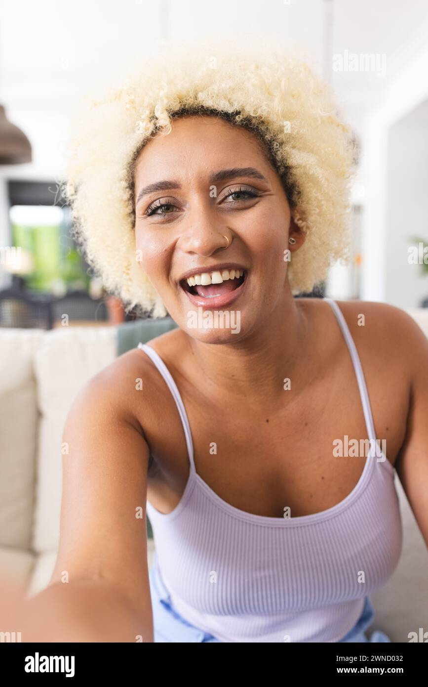 Jeune femme biraciale aux cheveux blonds bouclés sourit à la caméra lors d'un appel vidéo Banque D'Images