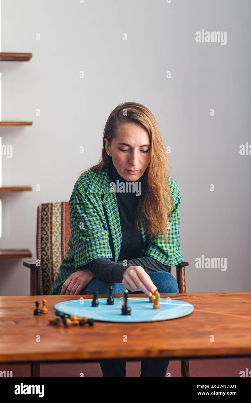 Composition verticale. Une femme déterminée s'assoit soigneusement à côté d'une table basse sur laquelle il y a des pièces d'échecs sur un cercle bleu et conduit un psyc Banque D'Images