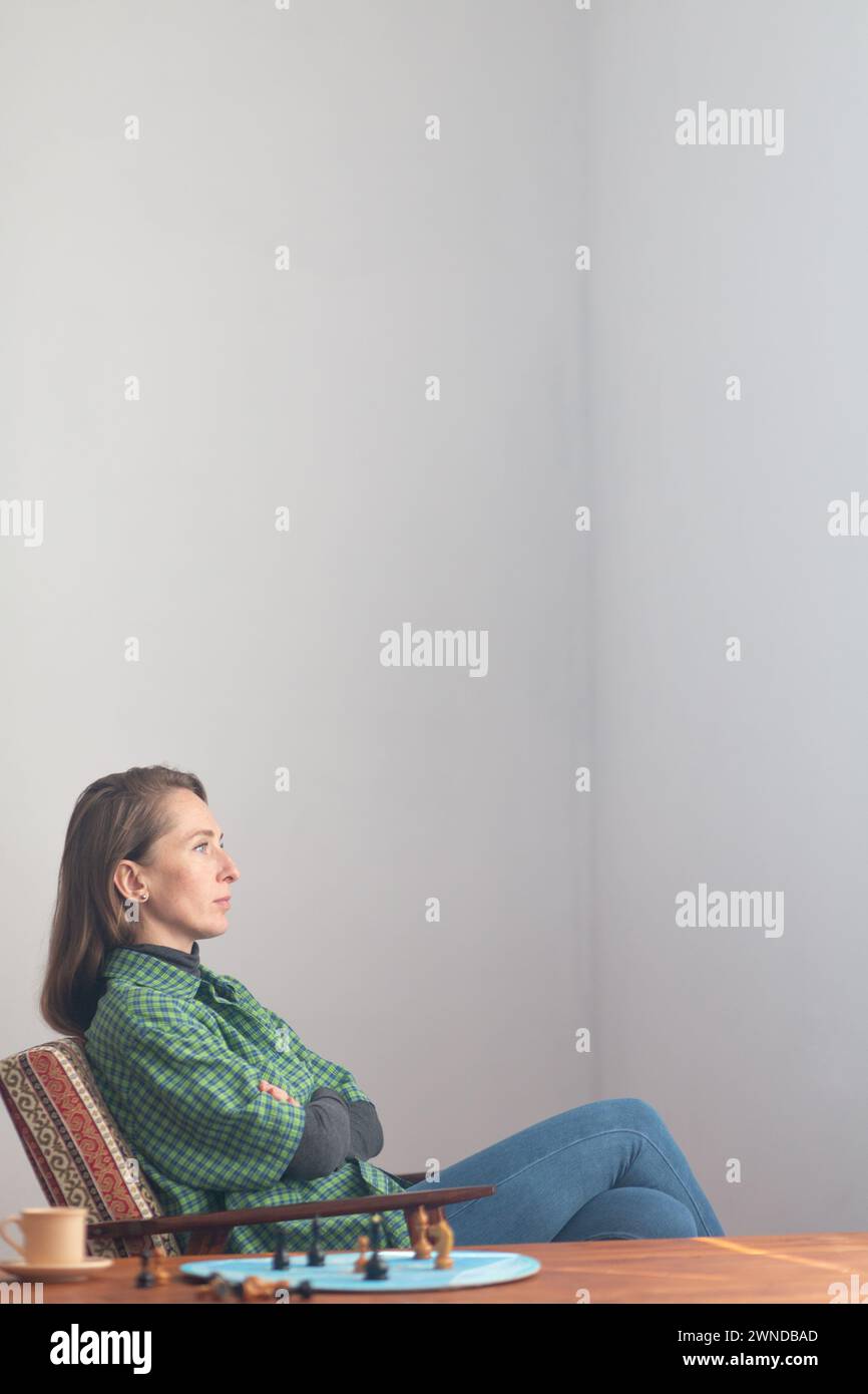 Composition verticale. Une femme déterminée est assise dans une chaise avec ses bras croisés sur sa poitrine et regarde attentivement devant elle. A proximité sur la table basse sont Banque D'Images