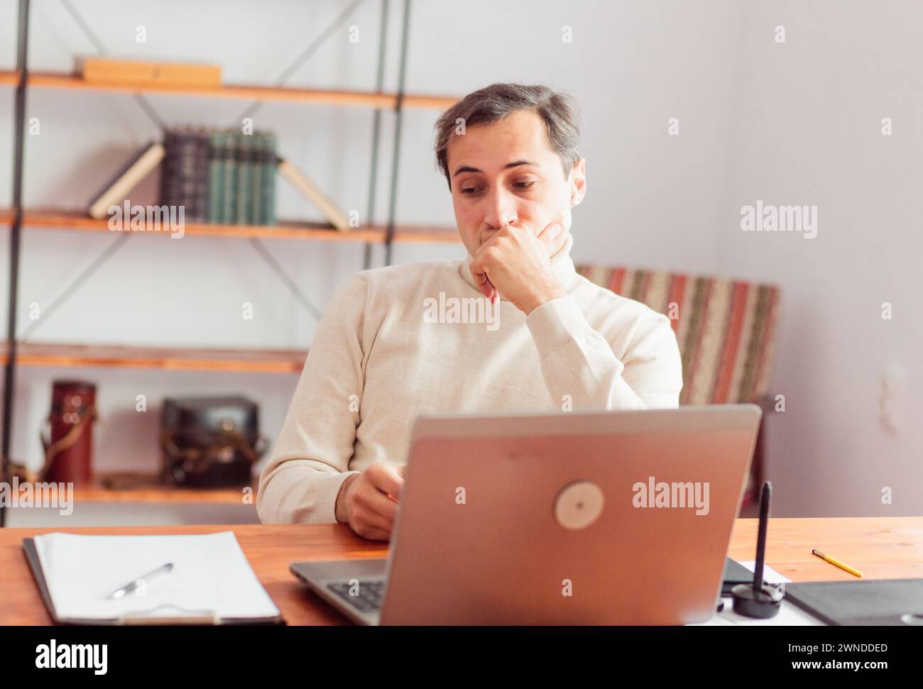 Un jeune homme adulte dans un bureau à domicile est réfléchi et regarde sur le côté avec sa tête appuyée sur sa main tout en travaillant sur un ordinateur portable et un téléphone portable Banque D'Images