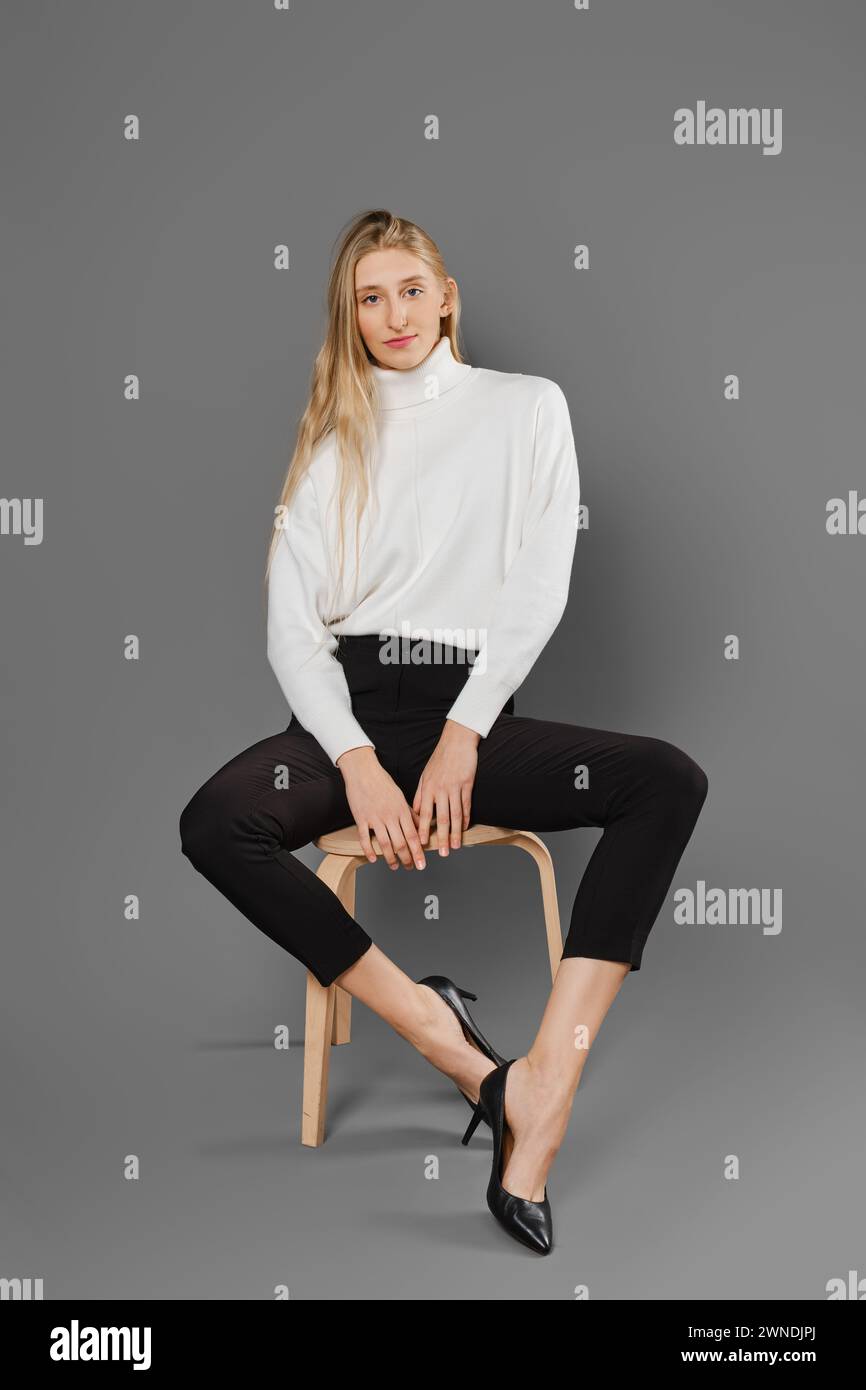 Jeune femme blonde en pantalon serré et pull est assis sur la chaise avec les jambes écartées sur les côtés Banque D'Images