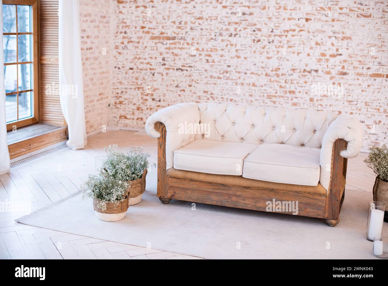 Un canapé matelassé blanc avec une base en bois devant un mur de briques blanches, à côté d'une grande fenêtre du sol au plafond avec des rideaux blancs et des pots de fleurs sur un Banque D'Images