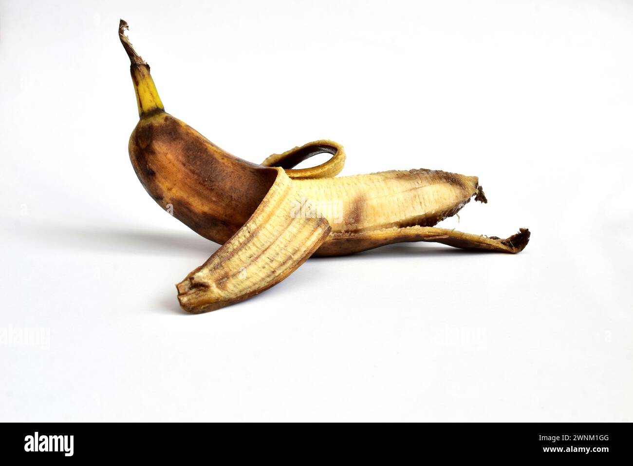 Sur un fond blanc se trouve une banane qui n'a pas été complètement pelée. La peau de banane a des taches sombres. La banane est trop mûre. Banque D'Images