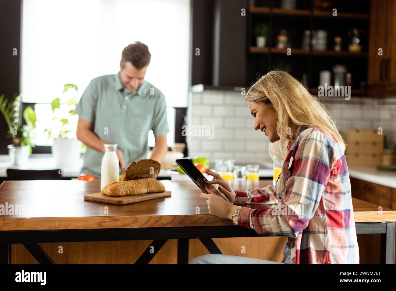 Une femme lit sur une tablette au comptoir de la cuisine tandis qu'un homme tenant une tomate lui sourit Banque D'Images