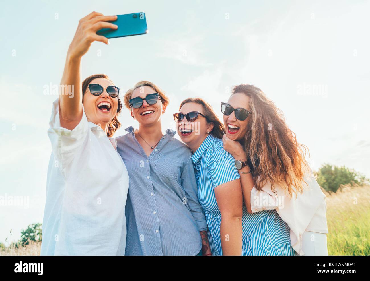 Portrait de quatre femmes souriantes et gaies dans des lunettes de soleil qui s'embrassent ensemble et font des photos de selfie à l'aide d'un smartphone moderne pendant la marche en extérieur. Femme Banque D'Images