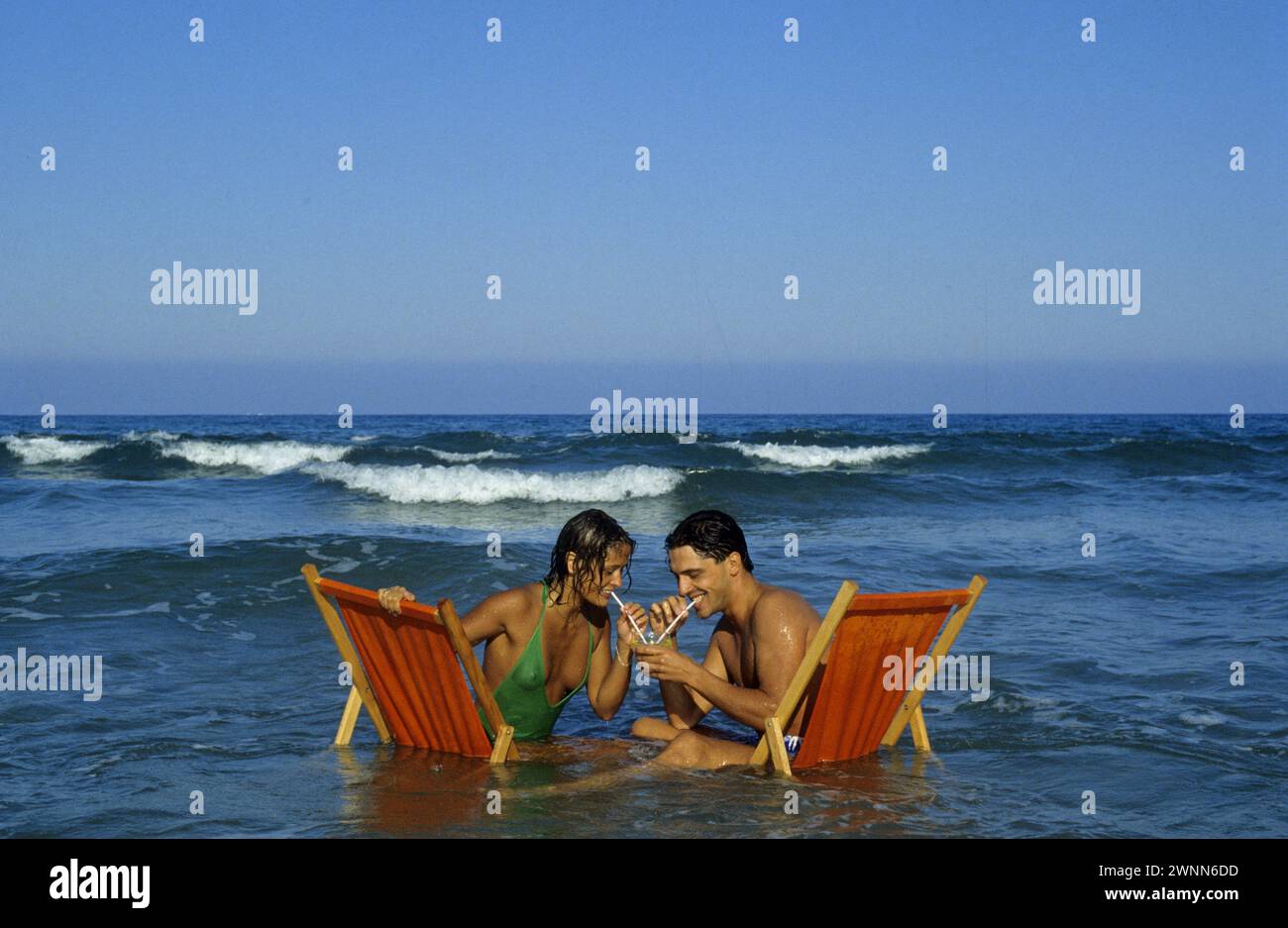 cheveux noirs jeune couple vacances d'été boire le même jus d'orange coktail au milieu des vagues insolite photographie drôle ambiance horizontale Banque D'Images