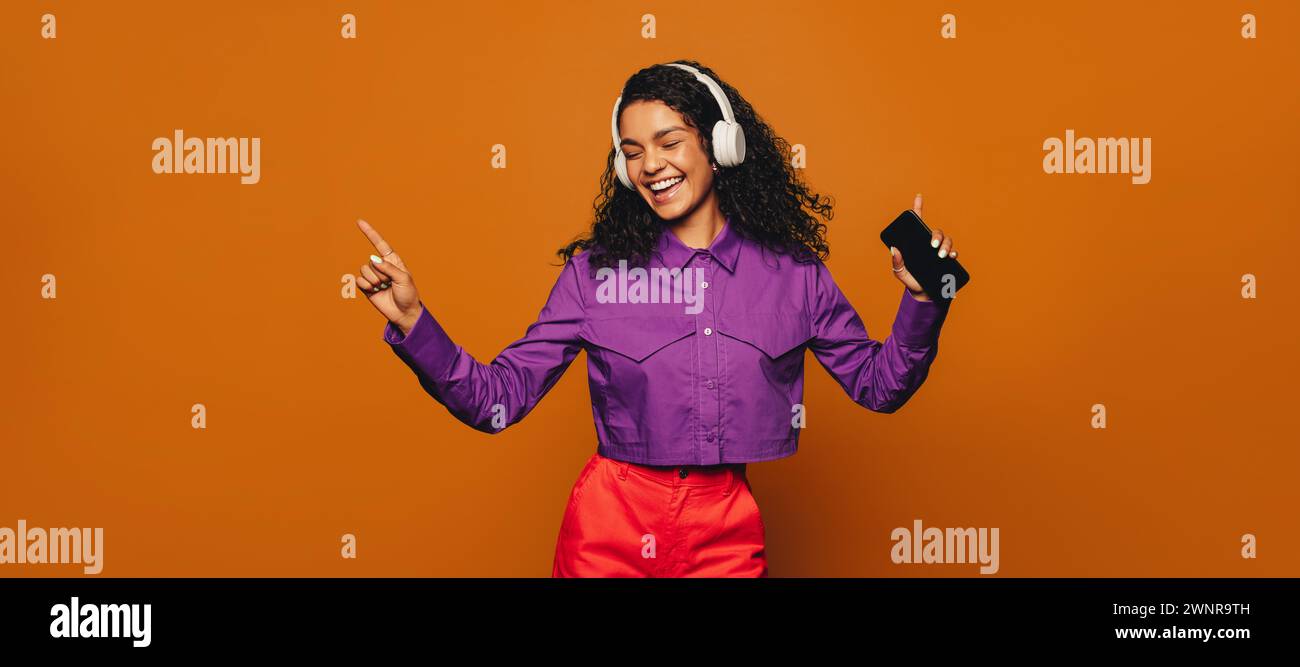 Femme joyeuse avec les cheveux bouclés se tient sur un fond orange vif, tenant un smartphone et portant des vêtements décontractés. Elle sourit comme elle danse et l Banque D'Images