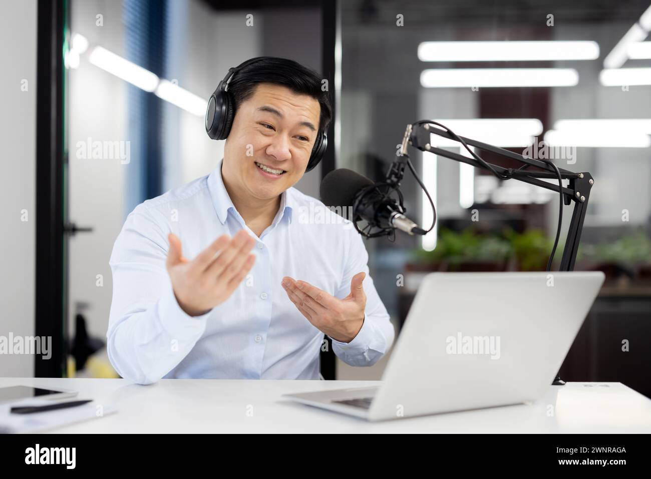 Souriant jeune homme asiatique portant des écouteurs assis dans le bureau devant le bureau avec microphone et parler sur appel vidéo sur ordinateur portable faisant des gestes avec les mains. Banque D'Images