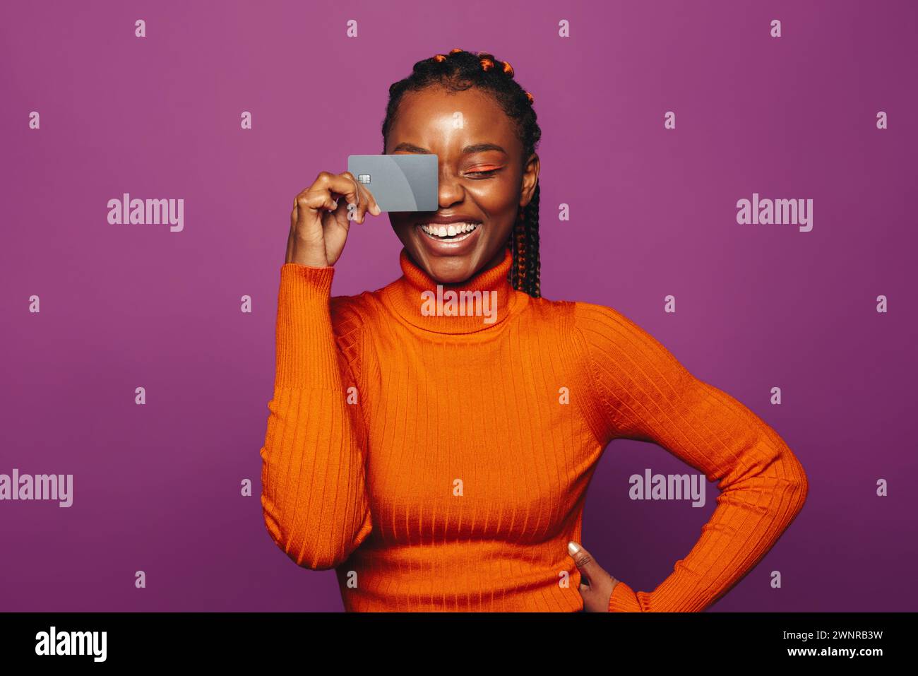 Jeune femme africaine avec des tresses se tient sur un fond violet coloré et détient une carte bancaire pour le paiement sans contact. Sa vie heureuse et décontractée Banque D'Images