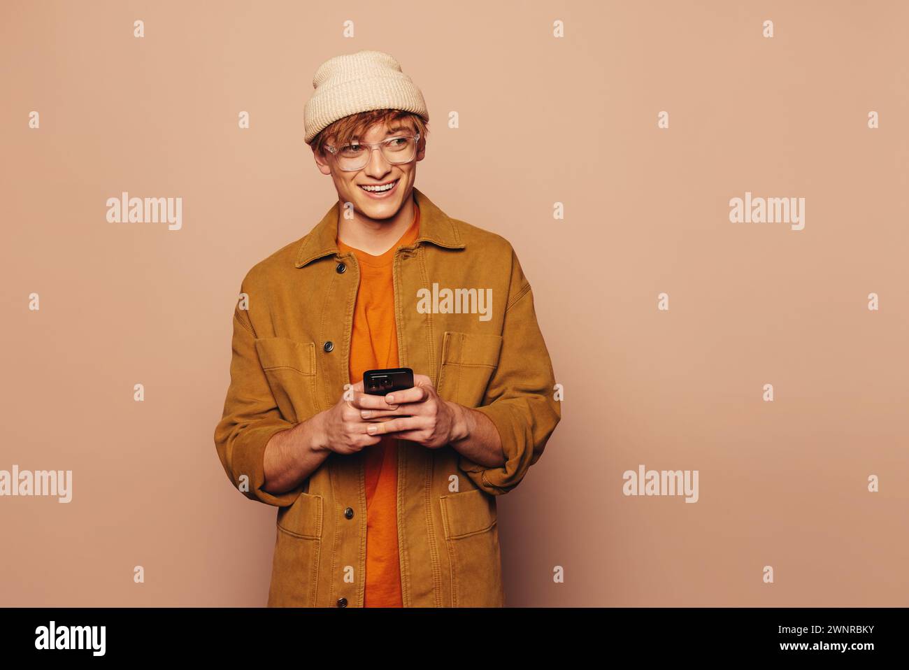 Portrait d'un jeune homme joyeux dans une veste en denim décontractée et des lunettes, heureusement à l'aide d'un téléphone portable. Fond pêche vibrant ajoute un moderne et colorfu Banque D'Images