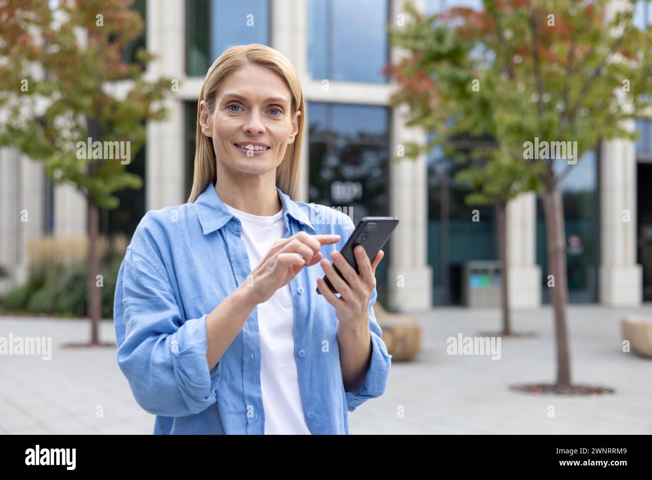 Une jeune femme adulte se tient dehors, concentrée sur son smartphone, avec des bâtiments modernes en arrière-plan. L'image capture une scène commune de connectivité et de style de vie urbain. Banque D'Images