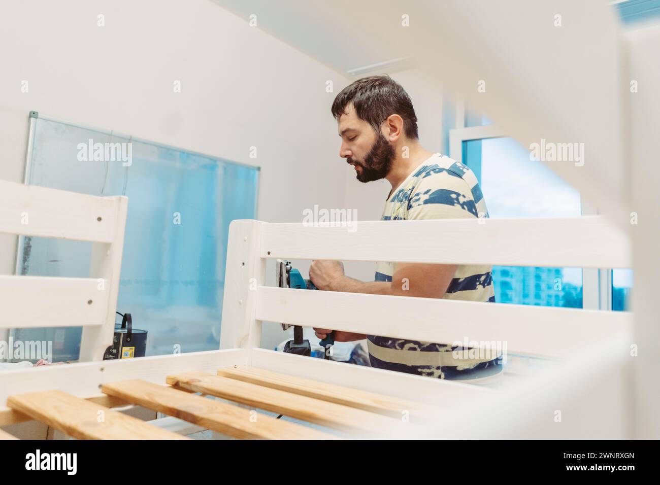 L'homme portant un masque de protection frotte le lit d'enfant en bois avec la ponceuse électrique ou le broyeur enlevant la vieille peinture.image avec la mise au point sélective Banque D'Images