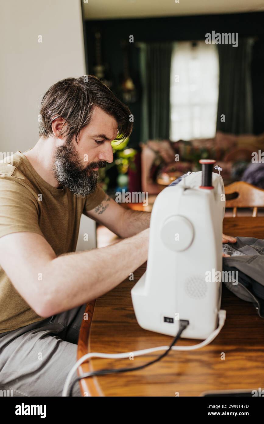 L'homme est assis à la table de la cuisine avec machine à coudre patching pantalon de travail Banque D'Images