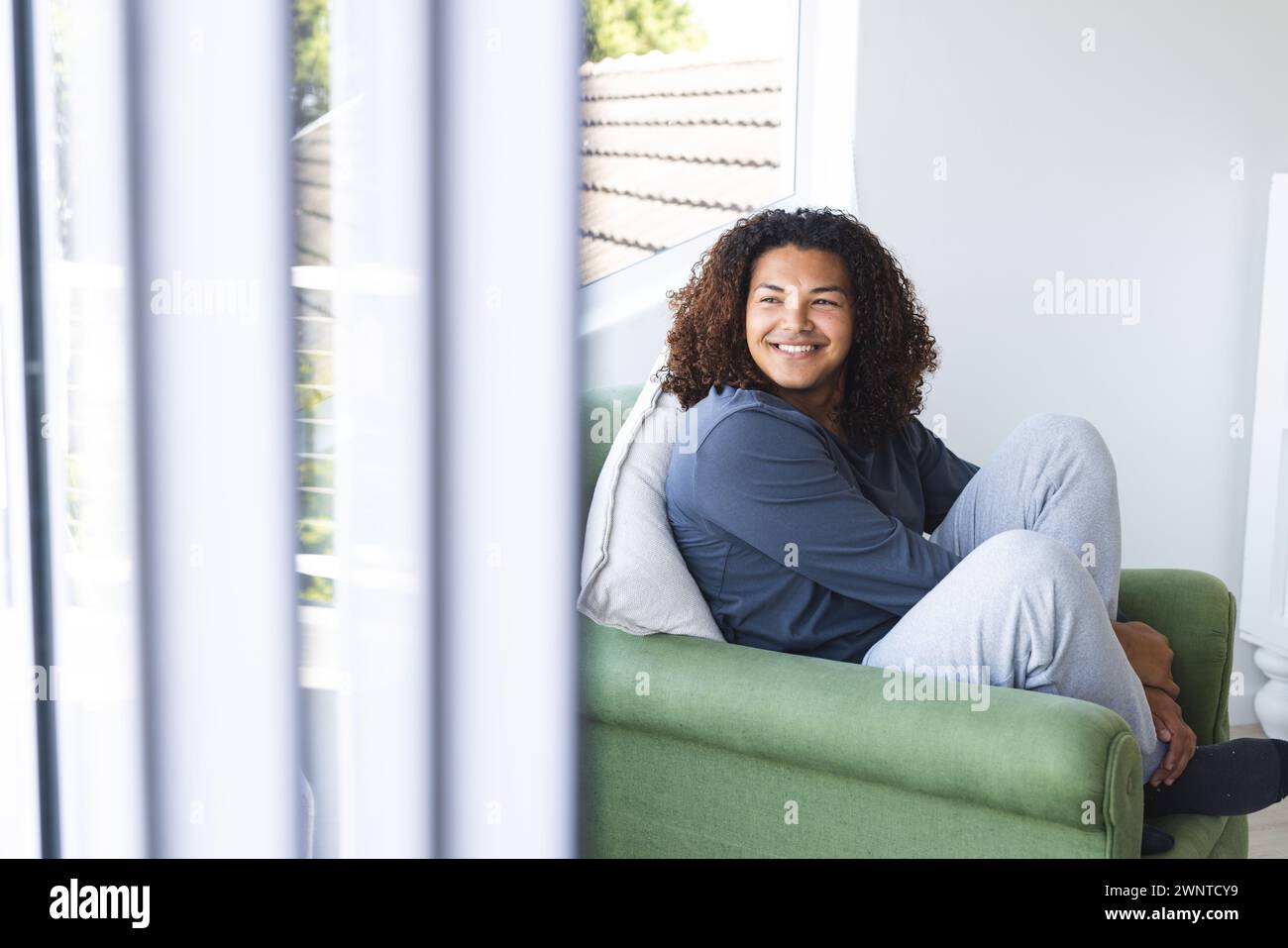 Jeune homme biracial avec les cheveux bouclés sourit tout en étant assis sur une chaise verte avec espace de copie Banque D'Images