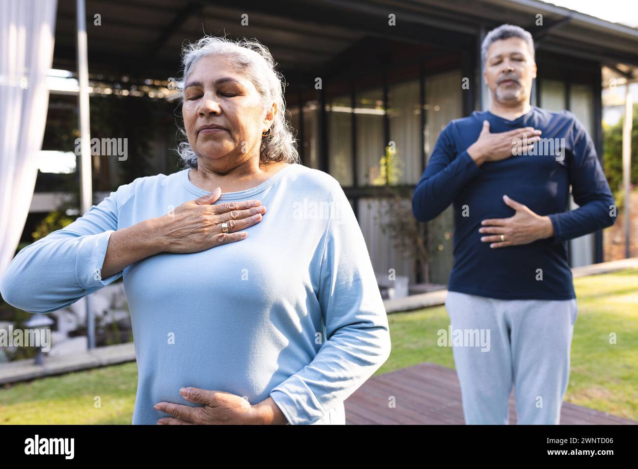 Femme biraciale senior et homme pratiquent le yoga à la maison, debout avec les yeux fermés et les mains sur les poitrines Banque D'Images