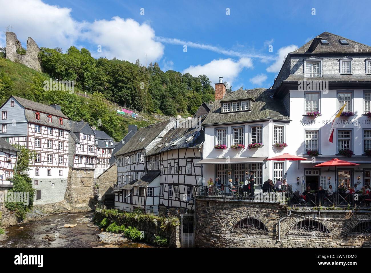 Pittoresque village européen (Monschau, Allemagne) avec des maisons à colombages et un pont fleuri sur un ruisseau, avec des collines verdoyantes en arrière-plan Banque D'Images