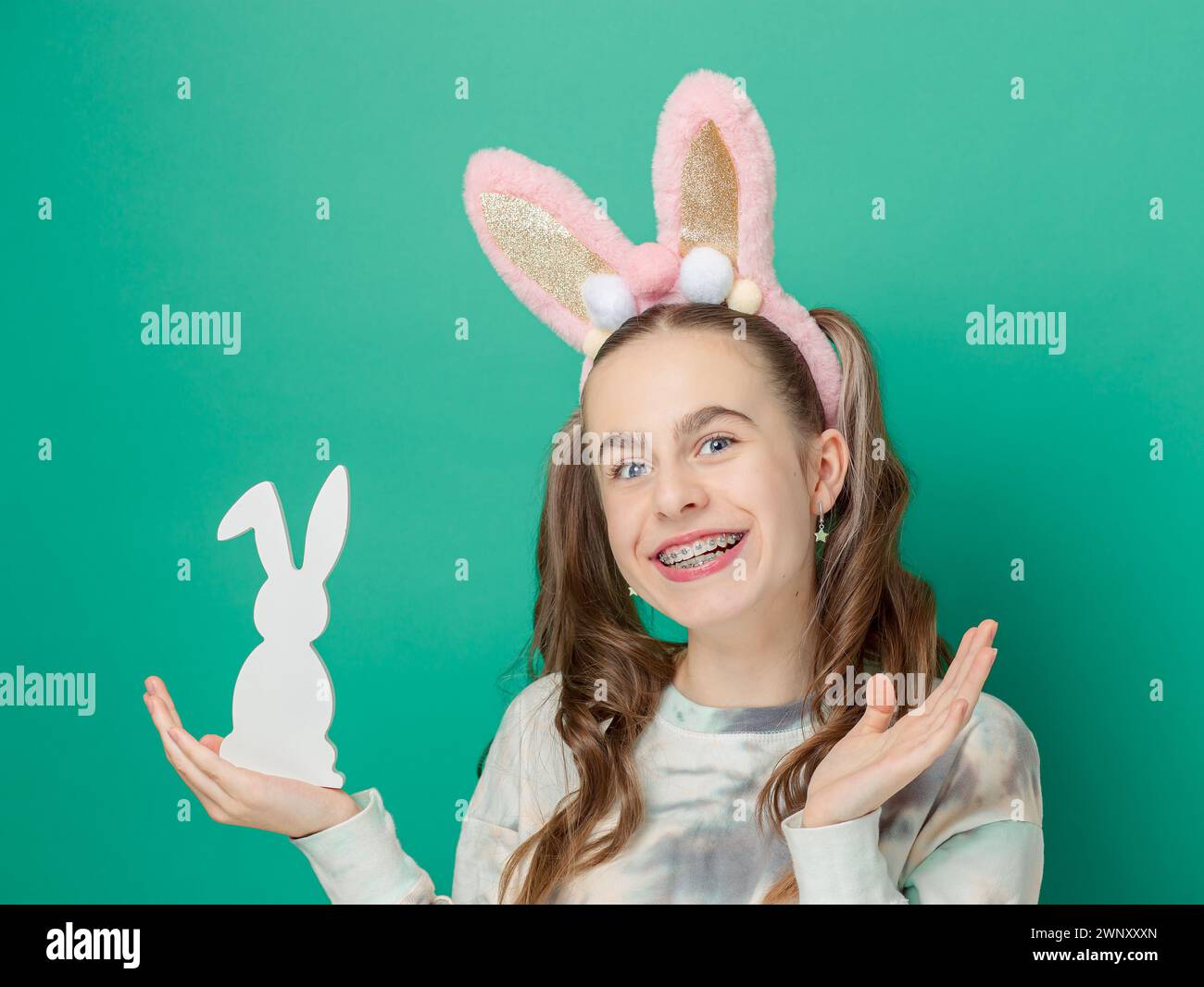 Joyeuses Pâques. Fille avec un sourire heureux, portant des oreilles de lapin rose brillantes et tenant une figurine de lapin en bois, fond bleu, rayonnant de plaisir de Pâques. Mignon smil Banque D'Images