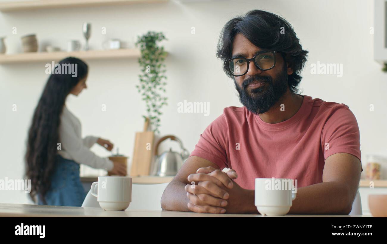 Heureux homme barbu mari propriétaire d'une maison assis dans la cuisine musulman indien souriant sourire joyeux tandis que femme femme cuisinant le petit déjeuner travail ménager culinaire Banque D'Images