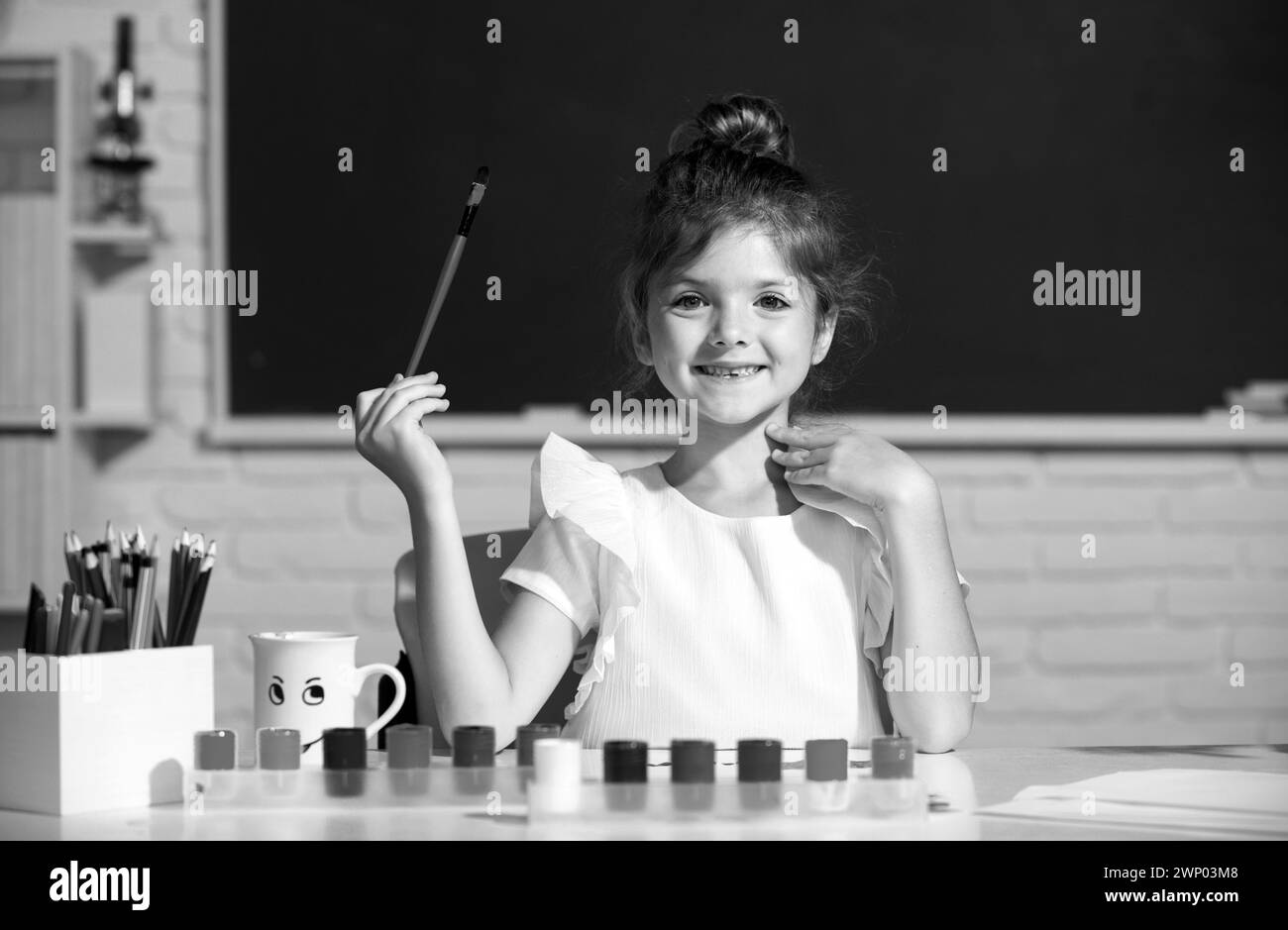 Croissance créative des enfants.Enfant fille tire dans la salle de classe assis à une table, en s'amusant sur fond de tableau noir de l'école. Banque D'Images