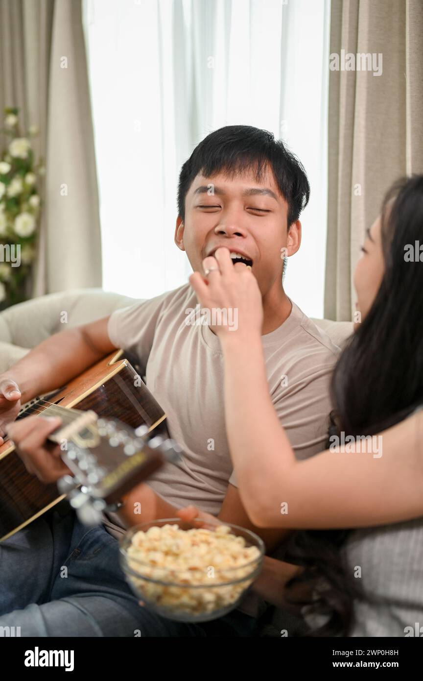 Une jeune petite amie attentionnée nourrit son petit ami de pop-corn pendant qu'il joue de la guitare, créant une atmosphère joyeuse ensemble à la maison. couple charmant, Banque D'Images