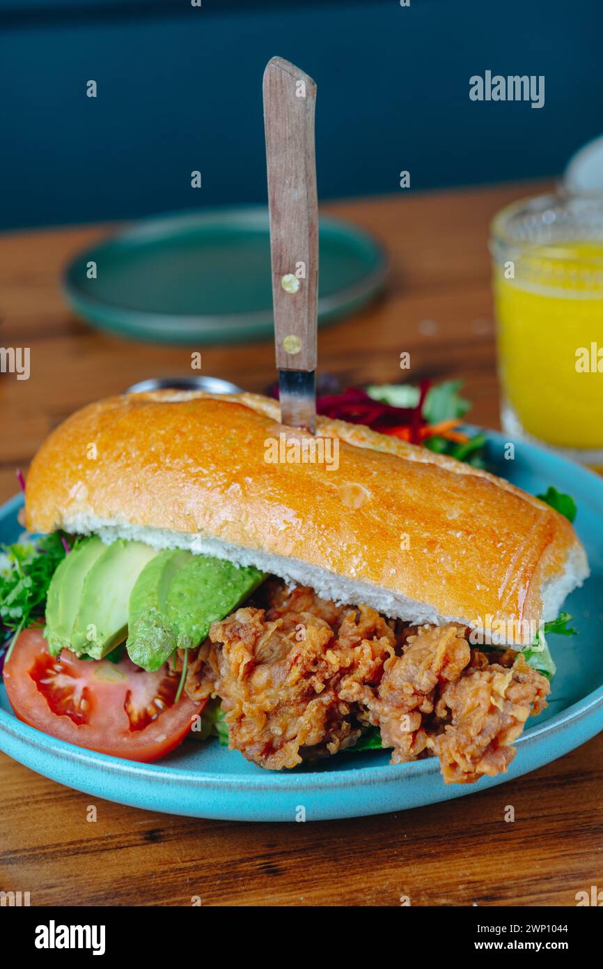 Un sandwich avec un couteau qui en sort est sur une plaque bleue. Le sandwich est garni de laitue, de tomates et d'avocat. Il y a aussi un verre de ou Banque D'Images