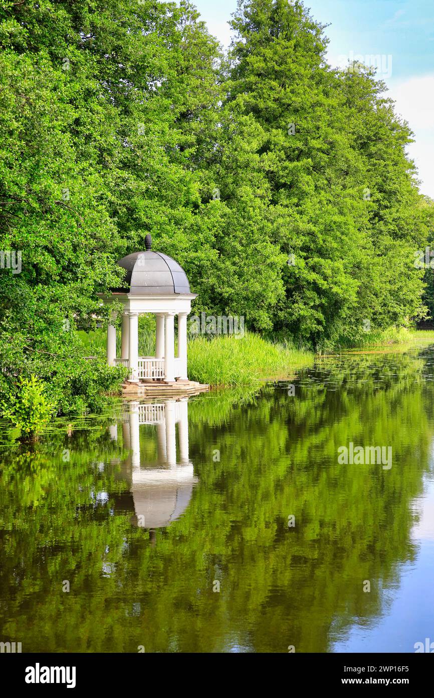 Belvédère blanc en bois au bord de la rivière entouré d'arbres. Vue sur un matin d'été paisible avec ciel clair et reflets sur l'eau. Banque D'Images