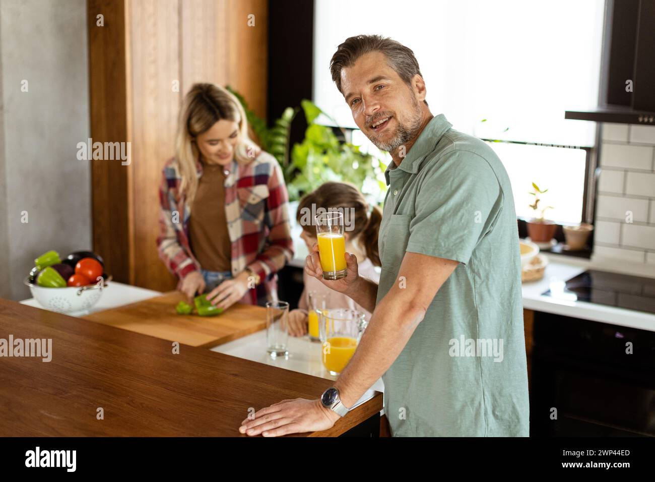 Un homme joyeux avec un verre de jus d'orange tandis qu'une femme et un enfant préparent la nourriture dans une cuisine confortable. Banque D'Images