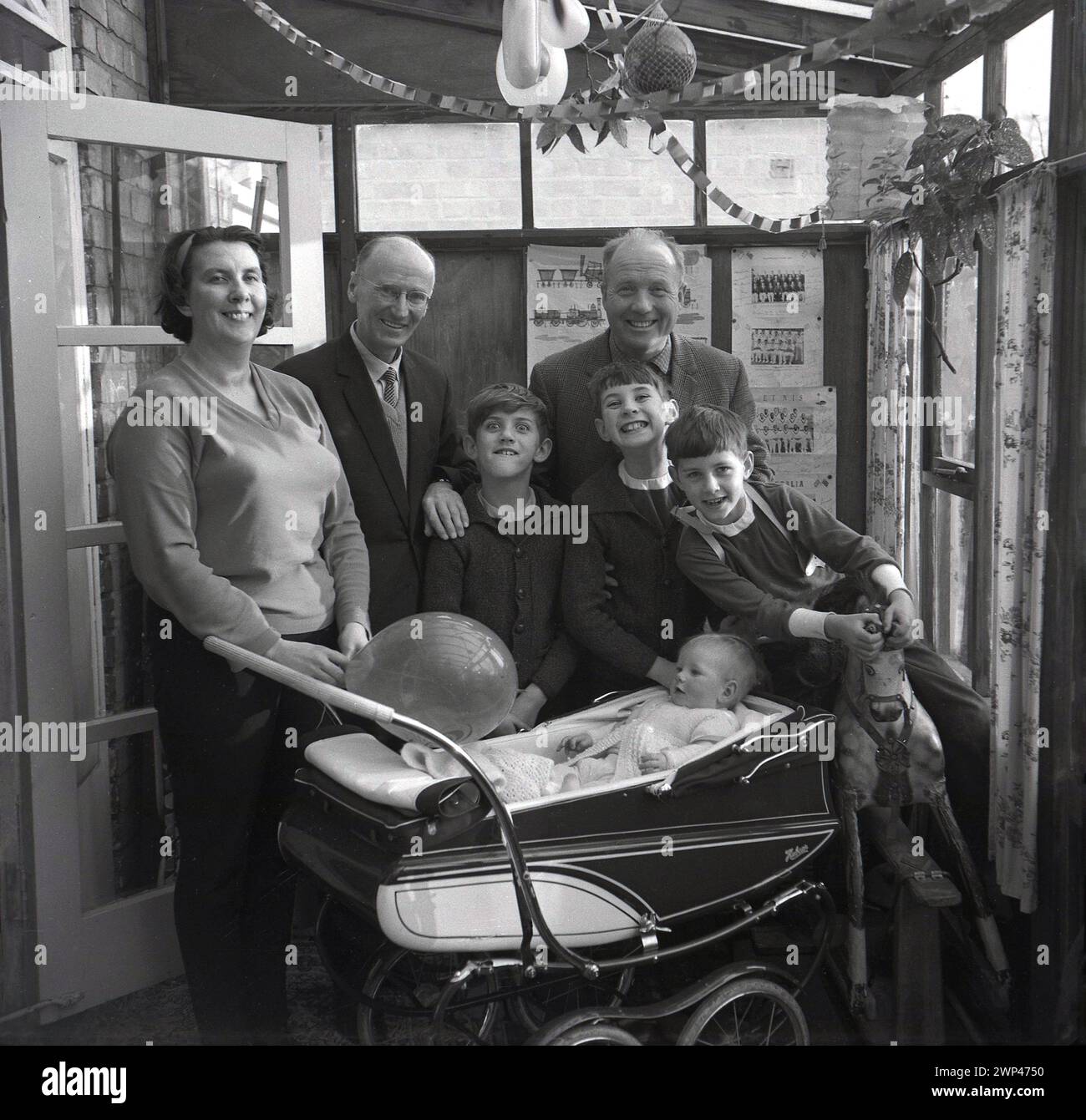 Années 1960, historique, grands sourires tout autour comme une famille debout pour une photo à l'intérieur d'un arrière-maison penché avec un enfant en bas âge assis dans une voiture d'entraînement construit tenant un ballon, Angleterre, Royaume-Uni. Banderole d'anniversaire au plafond. Banque D'Images