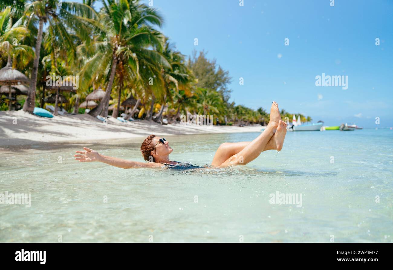 Belle femme brune habillée maillot de bain noir dans des lunettes de soleil de mode se relaxant allongé sur les vagues de plage de sable peu profondes avec des chaises longues de luxe exotiques, Banque D'Images