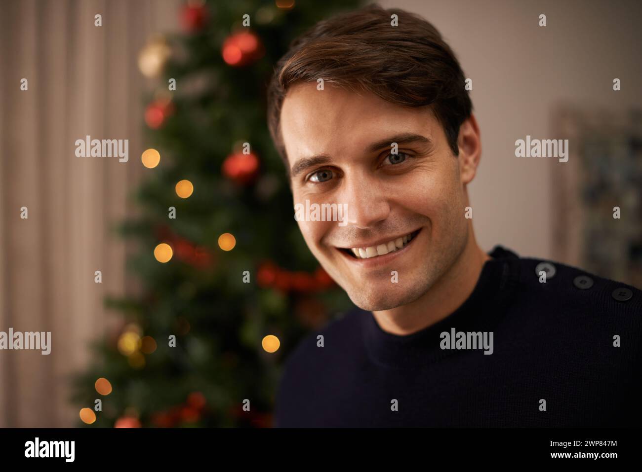 Portrait, maison et homme avec le sourire pour Noël dans le salon avec des décorations sur l'arbre pour la célébration. Appartement, bonheur et personne masculine avec joie Banque D'Images